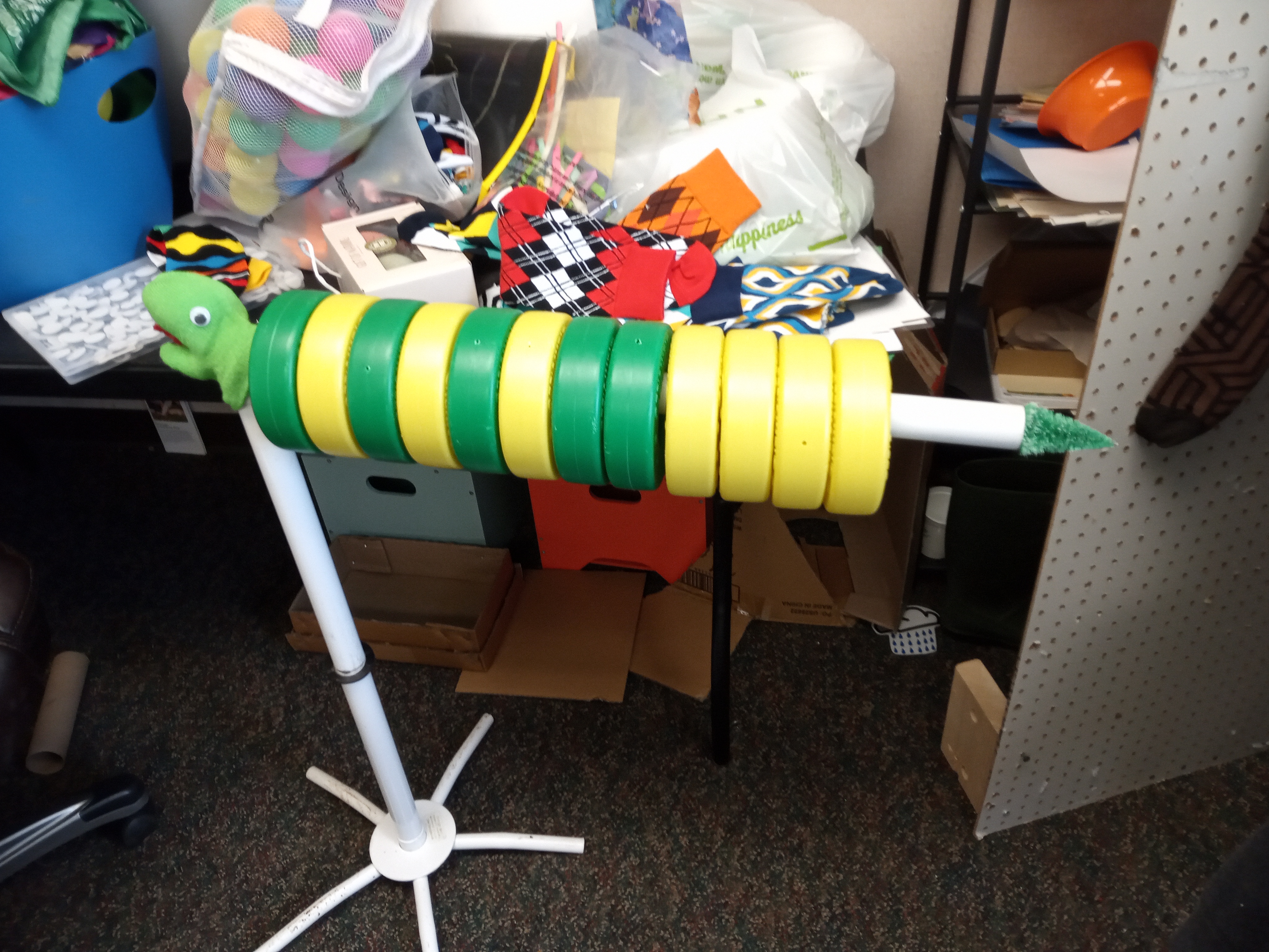 A white stand with a vaguely reptile looking green head with green and yellow disks lined up along its horizontal pipe.